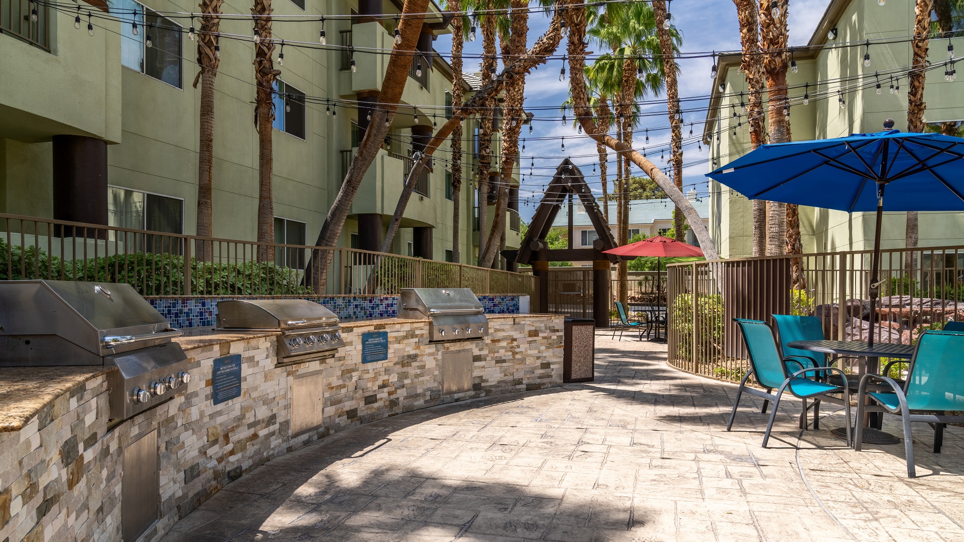 An outdoor BBQ area with stone countertops, stainless steel grills, and outdoor seating under blue umbrellas.