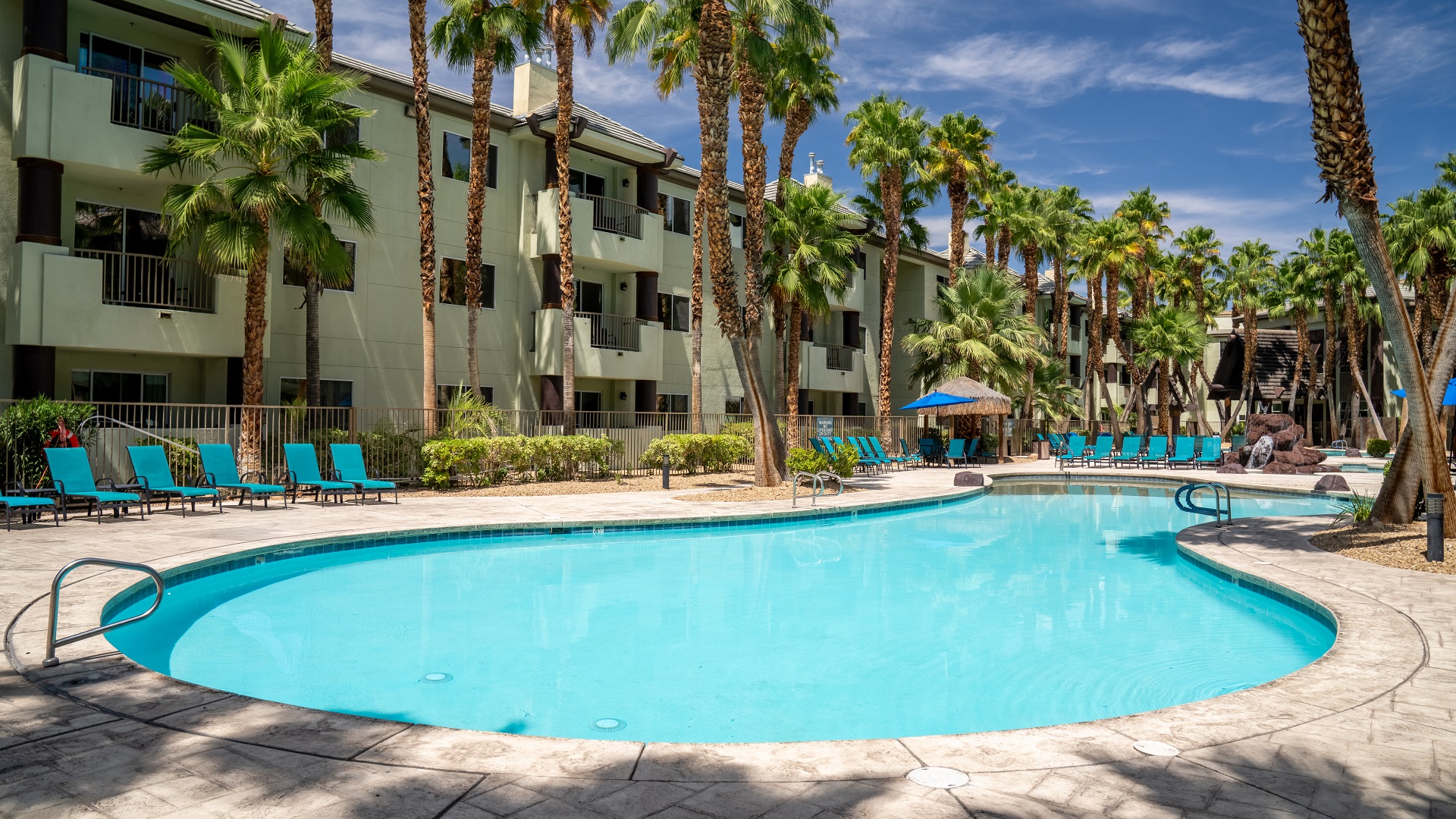 A large, kidney-shaped swimming pool with blue water, surrounded by beige tiles, blue lounge chairs, and palm trees, in front of a multi-story hotel building.
